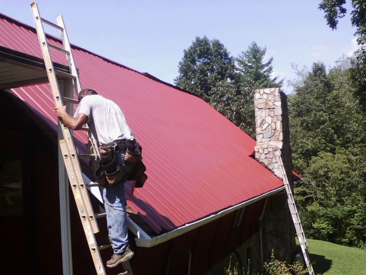 Roofer on a ladder beside a red metal roof and stone chimney — Soffit & Fascia Repair by Roofing Brothers in Sweetser